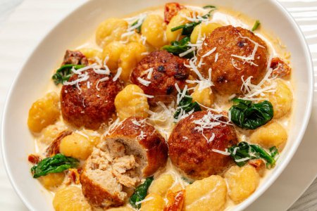 tuscan chicken meatballs with gnocchi in creamy sauce with sun-dried tomatoes and spinach leaves in white bowl on white wooden table, close-up
