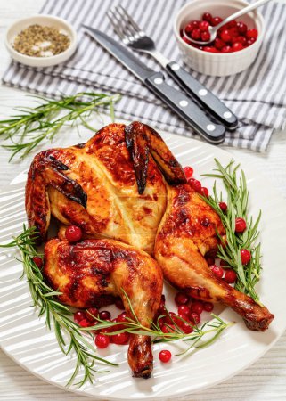 butterflied or spatchcock roast whole chicken on white plate with rosemary and cranberry on wooden table with cutlery and napkin, vertical view from above