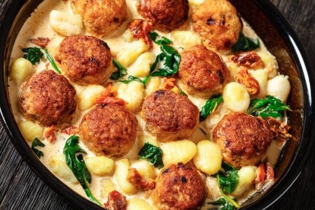 tuscan chicken meatballs with gnocchi in creamy sauce with sun-dried tomatoes and spinach leaves in baking dish on dark wooden table, close-up