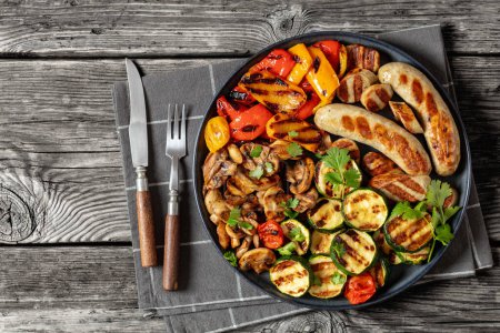 grilled white sausages Weisswurst with grilled peppers, zucchini and mushrooms on dark grey plate on rustic wooden table with fork, knife, horizontal view from above, flat lay, free space