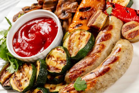 grilled white sausages Weisswurst, grilled yellow and red peppers, zucchini and mushrooms with ketchup on white plate, close-up