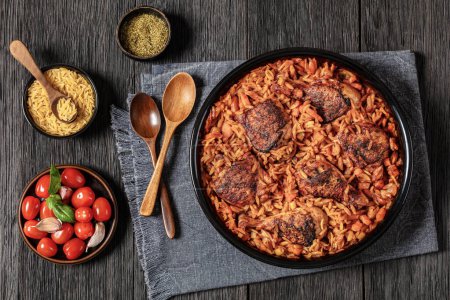 One pan baked greek chicken orzo stew in tomato based sauce, giouvetsi youvetsi in baking dish on dark oak wooden table with spoons and ingredients, horizontal view from above, flat lay