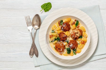tuscan chicken meatballs with gnocchi in creamy sauce with sun-dried tomatoes and spinach leaves in white bowl on white wooden table with spoon and fork, horizontal view from above, flat lay