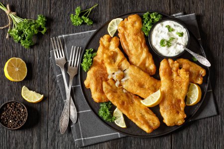 deep fried crispy beer batter cod fish fillet on black plate with tartar sauce and lemon slices on dark wooden table with forks, horizontal view from above, flat lay