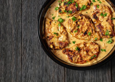 creamy smothered pork chops cooked in onion gravy in baking dish on dark oak wooden table, horizontal view from above, flat lay, close-up, free space