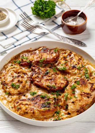 creamy smothered pork chops cooked in onion gravy in baking dish on white wooden table with fresh parsley, seasoning and forks, vertical view from above, close-up
