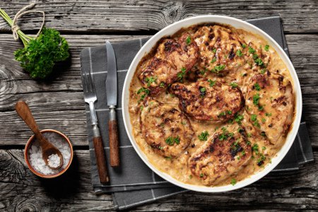creamy smothered pork chops cooked in onion gravy in baking dish on rustic wooden table with fresh parsley, salt and cutlery, horizontal view from above, flat lay