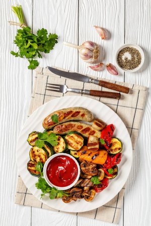 grilled white sausages Weisswurst with grilled peppers, zucchini and mushrooms on white plate on white wooden table with fork, knife, tomato sauce, vertical view