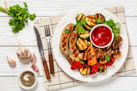 grilled white sausages Weisswurst, grilled yellow and red peppers, zucchini and mushrooms with tomato sauce on white plate on white  wooden table with cutlery, horizontal view from above, flat lay