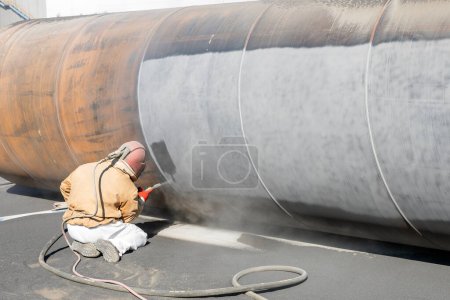 Téléchargez les photos : The sandblaster is sanding to steel pipe material. Abrasive blasting, more commonly known as sandblasting, is the operation of forcibly propelling a stream of abrasive material against a surface. - en image libre de droit