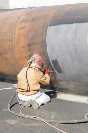 Téléchargez les photos : The sandblaster is sanding to steel pipe material. Abrasive blasting, more commonly known as sandblasting, is the operation of forcibly propelling a stream of abrasive material against a surface . - en image libre de droit