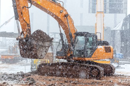 Téléchargez les photos : La grande excavatrice creuse par temps neigeux. Excavatrice dans la neige sur une route asphaltée. - en image libre de droit