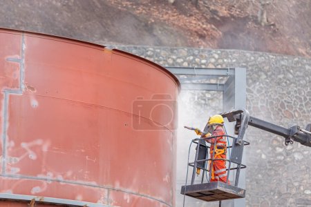 Der Sandstrahler ist Sandstrahlen (Abrasivstrahlen) t Stahlspeicher mit einem Hallo-ab-Kran auf der Baustelle.
