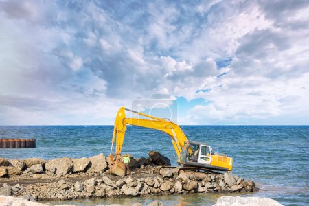 Téléchargez les photos : Travailleur réparer les pistes excavatrice par la mer. Les excavatrices sont un équipement de construction lourde composé d'une flèche, d'une benne (ou d'un bâton), d'un godet et d'une cabine en rotation. plate-forme. - en image libre de droit