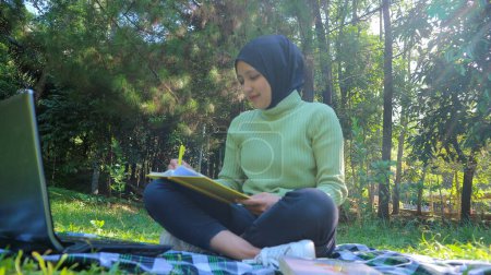 Foto de Relajada mujer musulmana disfrutando de fin de semana en el parque, sentado en la hierba y libro de escritura, espacio vacío - Imagen libre de derechos
