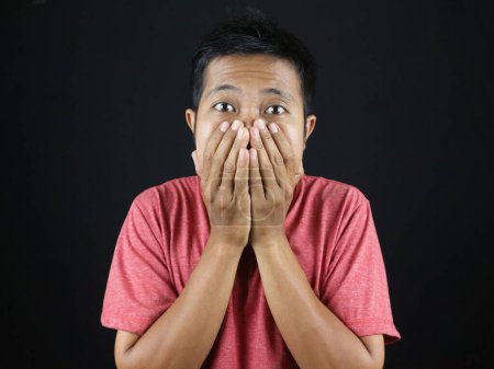 Young excited Asian man with hand covering mouth isolated on black studio background with copy space