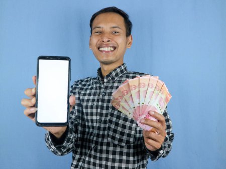 Foto de Emocionado expresión guapo asiático hombre celebrar billetes de papel moneda y mostrando pantalla de teléfono en blanco. Concepto de negocio y finanzas. - Imagen libre de derechos