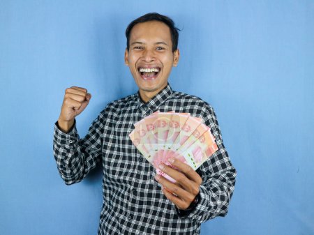 Excited expression handsome asian man hold paper money banknotes with clenched fist gesture. Business and finance concept.
