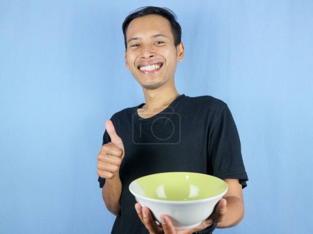 Ein junger asiatischer Mann im schwarzen T-Shirt steht auf und hält eine leere Schüssel mit dem Teller in der Hand.