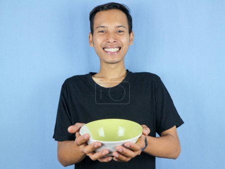 Ein junger asiatischer Mann im schwarzen T-Shirt steht auf und hält eine leere Schüssel mit dem Teller in der Hand.