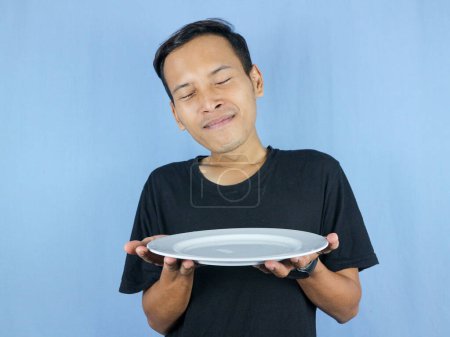 young Asian man in a black t-shirt stands and holds an empty white plate with the gesture of inhaling the dish.
