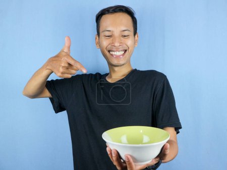 A young Asian man in a black t-shirt stands and holds an empty bowl showing the dish.