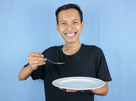 A young Asian man in a black t-shirt stands and holds a spoon and empty white plate with the gesture of preparing to devour the dish.