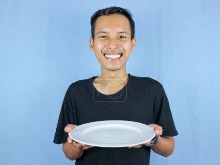 Ein junger asiatischer Mann im schwarzen T-Shirt steht auf und hält einen Löffel und einen leeren weißen Teller mit der Geste, sich vorzubereiten, das Gericht zu verschlingen.