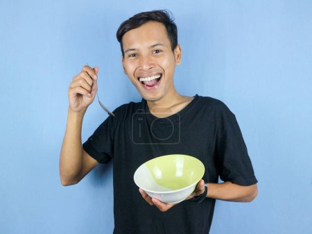 Un jeune Asiatique en t-shirt noir tient une cuillère et un bol vide avec le geste de se préparer à dévorer le plat.