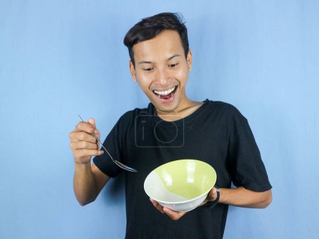Ein junger asiatischer Mann in einem schwarzen T-Shirt steht auf und hält einen Löffel und eine leere Schüssel mit der Geste, das Gericht zu verschlingen..