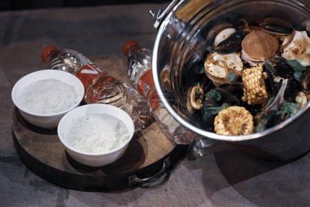 Portrait of a mixed seafood menu containing various shellfish, corn, etc. in a stainless bucket with rice in a white bowl and mineral water served on a wooden table.
