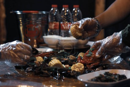 Großaufnahme von Händen mit Plastikhandschuhen, die Krabben auf einem gemischten Fischmenü aufheben, das auf einem Holztisch mit drei Schalen Reis und Mineralflaschen serviert wird.