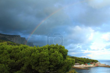 Foto de Rainbow in the coniferous park against the background of the mountains after a summer rain with bloated clouds - Imagen libre de derechos