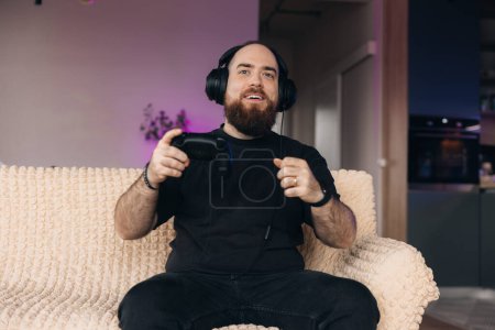 Téléchargez les photos : Joueur masculin jouant à des jeux vidéo avec le contrôleur dans ses mains et ses casques. Expression faciale d'un joueur. - en image libre de droit