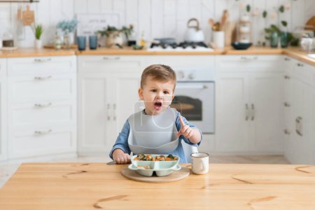 Photo for Cute caucasian toddler boy eats broccoli on his own using a fork. Self-feeding concept. BLW. The child eats healthy vegetables with meat on a high chair in the cozy kitchen. Copy space, Mock up. - Royalty Free Image