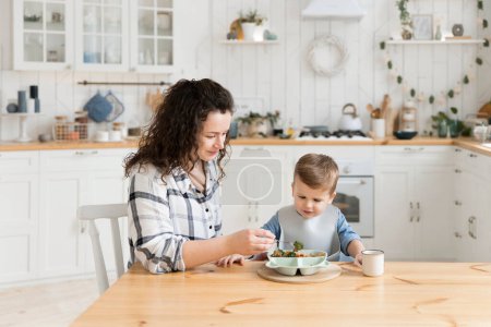 Foto de Young caucasian mother feeds her adorable toddler son with healthy vegetables and a chicken cutlet in a bright cozy scandinavian style kitchen. Childs lunch. Mock up, copy space. High quality photo - Imagen libre de derechos
