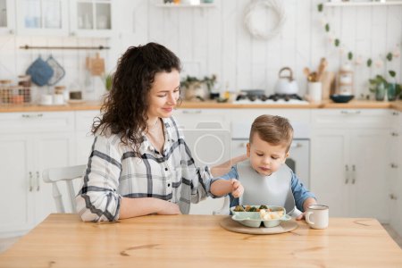 Photo for Young woman watches with delight as her son learns to eat on his own with a fork. Beautiful caucasian woman practices self-feeding with her baby. BLV. Dinner in a light and cozy kitchen. Copy space. - Royalty Free Image