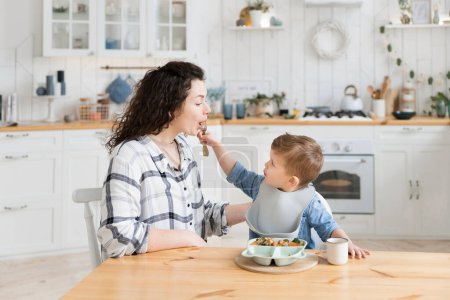 Photo for Adorable toddler boy feeding his young mother while she tries to feed him healthy vegetables and chicken cutlet. The child refuses to eat broocoli and gives it to his mother. Mock up, copy space. - Royalty Free Image
