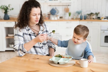 Foto de Adorable niño se niega a comer verduras que su madre le ofrece. Una madre joven se preocupa de que su hijo no quiere comer verduras y trata de persuadirlo de comer un pedazo de brócoli. - Imagen libre de derechos