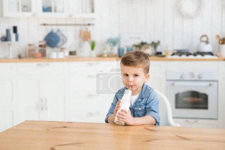 Photo for Cute baby toddler drinks milk on a blurred background of a light cozy kitchen. Handsome caucasian boy drinks vitamin rich organic lactose free yogurt with a straw. Copy space, product mock up. Tasty. - Royalty Free Image