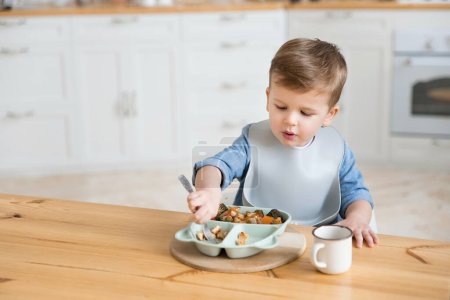 Photo for Adorable caucasian toddler boy eats vegetables on his own, pricking them on fork. The concept of self-feeding. BLW. Child eats healthy vegetables with meat on a high chair. Mock up. Place for text. - Royalty Free Image
