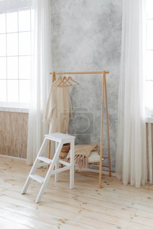 Photo for Stylish wooden coat rack and white wooden ladder stool in a spacious interior with large windows with curtains. Nobody. Wooden floor in the apartment. - Royalty Free Image