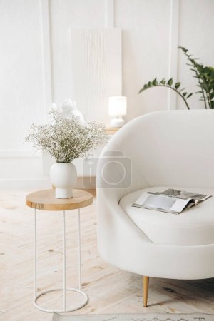 Photo for A stylish white leather sofa in a white living room and a minimalist coffee table with a bouquet of gypsophila in a white vase against the backdrop of a potted plant. Nobody. - Royalty Free Image