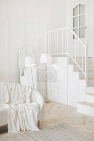 Photo for Stylish white living room with white leather sofa, floor lamp, full length oval mirror and wooden stairs leading to second floor. Staircase in appartament. Nobody. - Royalty Free Image