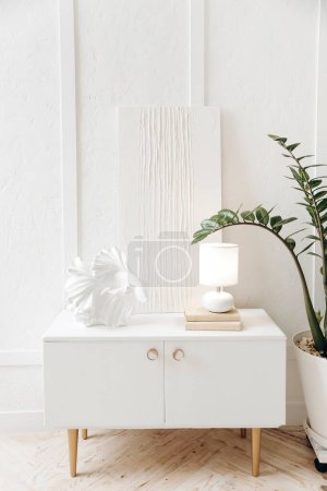 Téléchargez les photos : Table de chevet élégante blanche avec décor, lampe et peinture abstraite de gypse blanc contre un mur blanc texturé. Plante en pot à la maison. Personne.. - en image libre de droit