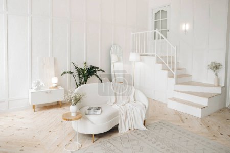 Photo for Stylish white living room with white leather sofa, floor lamp, full length oval mirror and wooden stairs leading to second floor. Staircase in appartament. Potted plant at home. Nobody. - Royalty Free Image