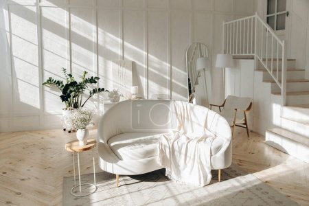 Téléchargez les photos : Élégant salon blanc avec canapé en cuir blanc, lampadaire, miroir ovale pleine longueur et escalier en bois menant au deuxième étage. Escalier. Plante en pot à la maison. Rayons de soleil à l'intérieur. Personne.. - en image libre de droit