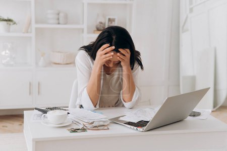 Desperate young woman holds head in hands, feeling overwhelmed by financial paperwork at white office desk, struggling with bills, taxes, and credit management, bankruptcy concept, mental health.