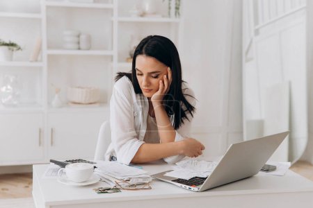 Tired young brunette woman at white office desk with laptop, visibly worried while sifting through financial papers, highlighting challenges of tax errors, ideal for financial advisory services.