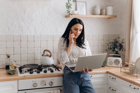 Confident young woman freelancer successfully balancing remote work and home chores, talk on the phone call and work on laptop in a well-organized, stylish kitchen. Concept of lifestyle management.